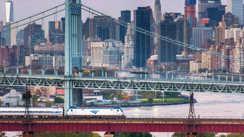 Silver train on bridge passing in front of green car bridge and tall colorful buildings.